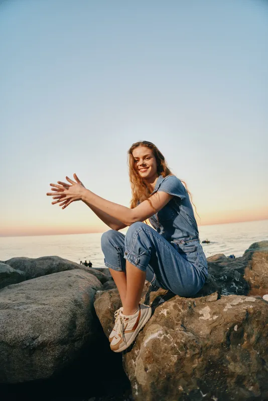 Lifestyle woman in modern boho-western denim street style sitting on rocks near water, smiling happily in film color warm sunset light, casual and free-spirited vibe.