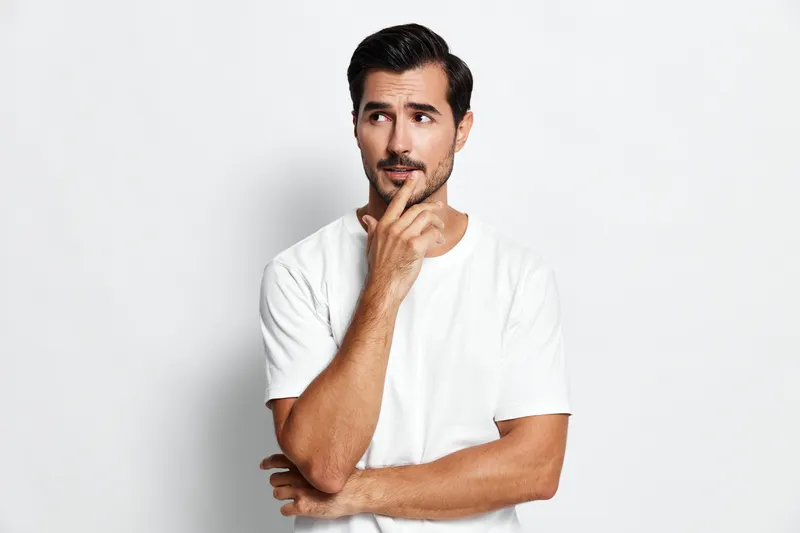 Thoughtful man in casual white t-shirt looking upwards with a pensive expression, hand on chin, isolated on plain light gray background. Emotion contemplation concept