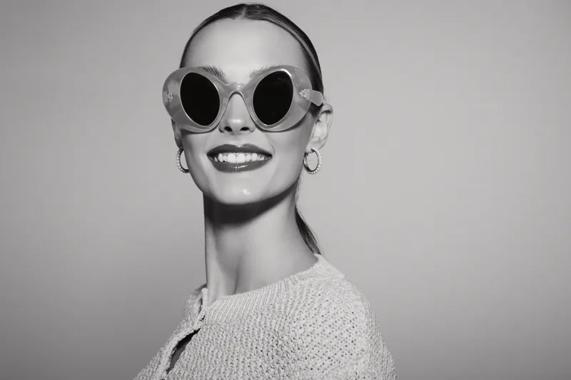 Smiling young Caucasian woman with fair skin wearing stylish sunglasses and earrings poses confidently against a plain background in a bright studio setting.