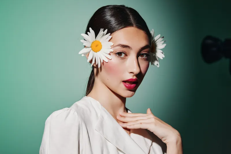 Young woman with smooth skin wearing white blouse poses with daisies in hair against a green background. Creative fashion beauty portrait with a fresh and natural vibe.
