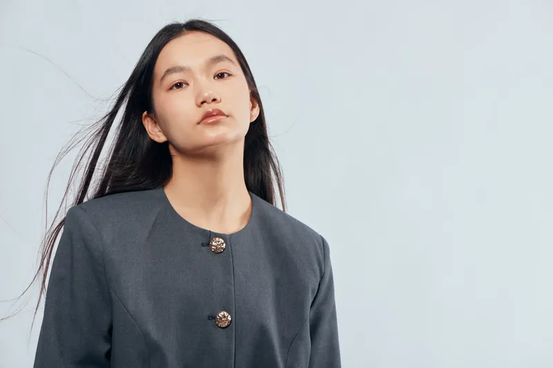 Young woman with long dark hair and grey jacket standing against light background looking confident and serious with natural expression in studio portrait.