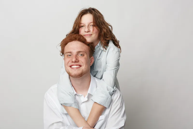 Smiling young couple in white shirts embraces and poses against plain background. Happy man and woman show affection and joy in casual studio portrait. Concept of love and friendship.