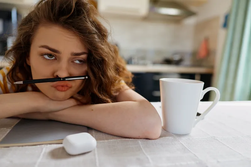 A tired woman sits at a home desk with a pencil in her mouth, a mug beside a notebook, and soft daylight on the table. A realistic daytime moment of focus, routine, and cozy concentration.