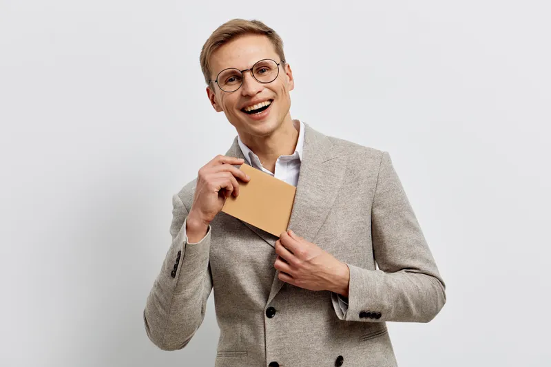 Confident smiling mature caucasian man with glasses in beige blazer holding small envelope against plain white background studio portrait. People lifestyle concept