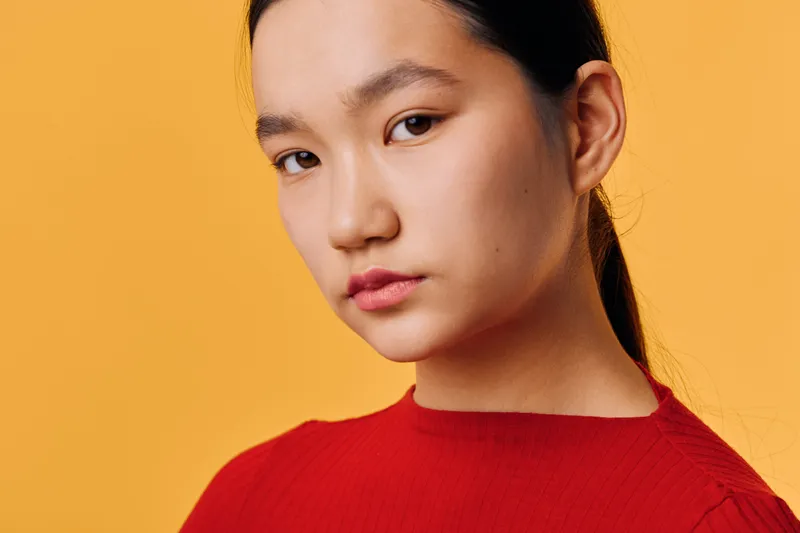 Portrait of young woman with long hair in red sweater looking at camera with serious expression on orange background indoors. Closeup, natural light, casual style and confident mood.