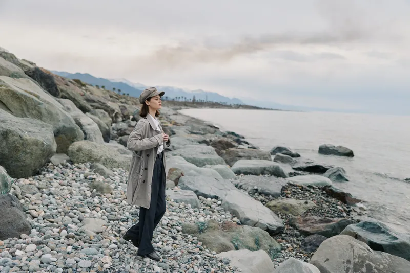 Woman in coat and hat stands on rocky beach holding camera, looking into distance. Overcast sky and calm sea create peaceful atmosphere. Concept of travel and nature exploration.