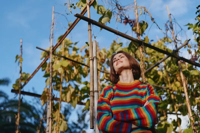 Woman in colorful sweater portrait outdoors in garden among bamboo poles, smiling and looking up in warm sunlight, casual cozy fashion, natural plants and relaxed pose.