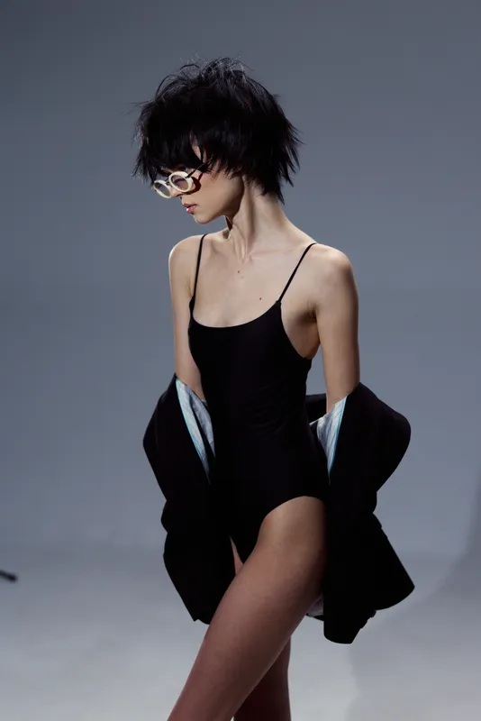 slim woman in black bodysuit wearing sunglasses with messy short hair against gray background