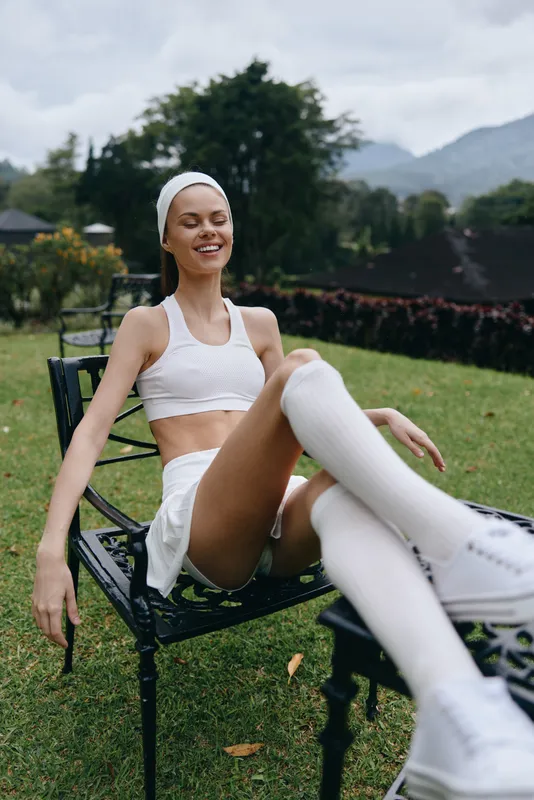 Smiling athletic woman in sportswear, relaxing in a lush garden, embodying fitness and wellness with a serene background