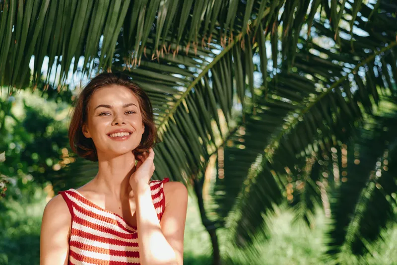 A cheerful woman stands outdoors in a sunny garden, wearing a striped tank top. She smiles warmly, enjoying the fresh air and natural light, creating a lively, relaxed mood for lifestyle and travel