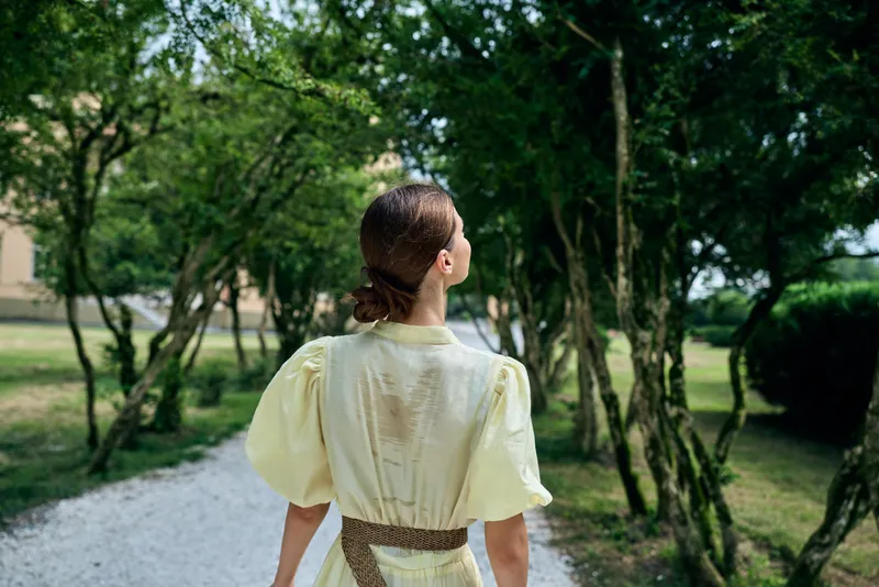 Woman in a light yellow dress walks along a garden path outdoors among trees, creating a serene nature scene with warm light and calm atmosphere.