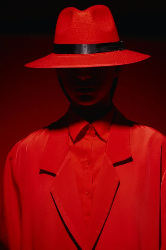 Close-up of a mysterious fashion model dressed in vibrant red clothing and a wide-brimmed hat, standing against a deep red background. Concept of beauty, style, and creative fashion.