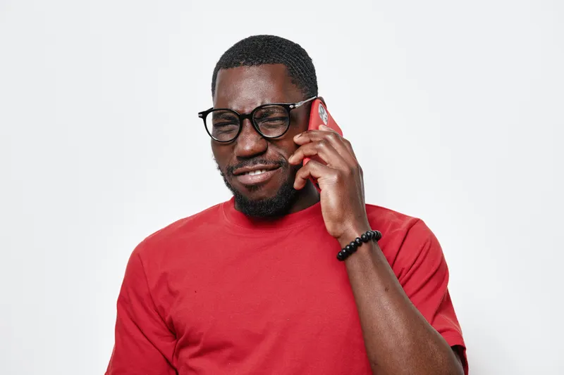 African American man with dark skin wearing glasses and red shirt talking on smartphone with smile isolated on white background. Casual communication and technology concept.