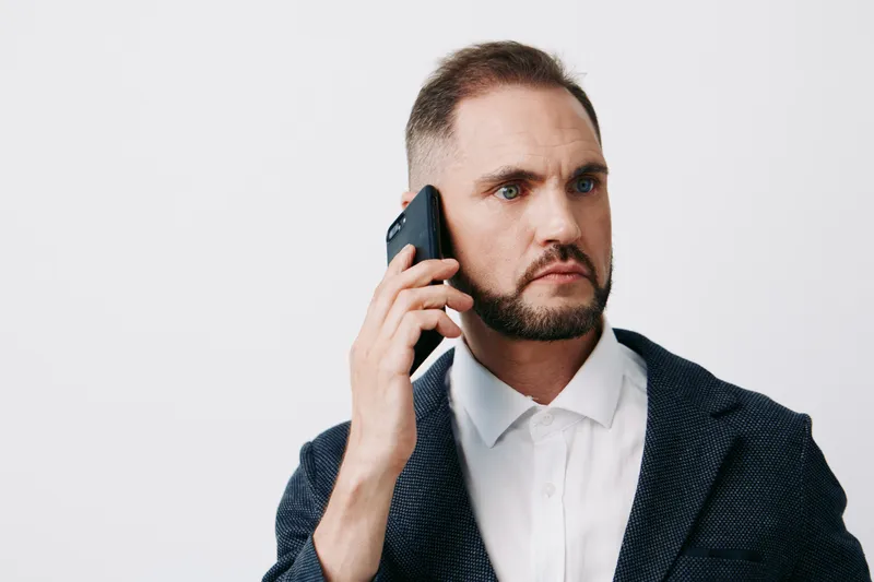 A focused business professional man on a solid colored background, talking on a mobile phone with a serious expression, conveying confidence, determination, and urgency.