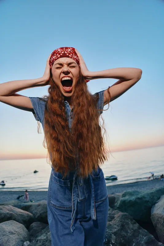 Lifestyle woman wearing modern boho-western denim street style screaming with hands on head at seaside in film color tones during sunset.