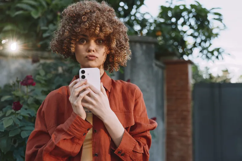 Cheerful woman outdoors, curly hair, taking a selfie, warm sunset glow, casual jacket, relaxed lifestyle moment, natural light, green backdrop