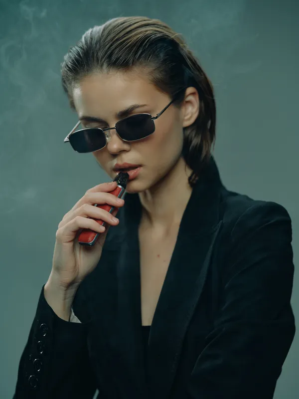 Stylish woman vaping with sunglasses indoors wearing black clothing and a confident expression