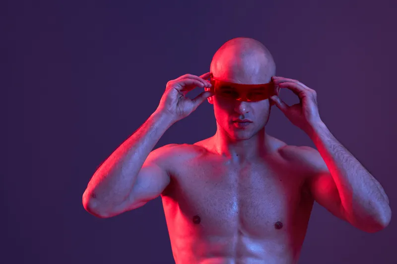 A confident man with a muscular build wearing futuristic sunglasses, showcasing a stylish look against a creatively lit backdrop in vibrant colors.