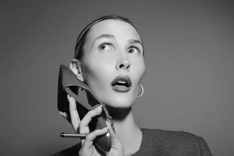 Young Caucasian woman with pale skin holds red high heel near face, looking up with surprised expression. Concept of fashion, beauty, elegance, and modern style in studio monochrome.