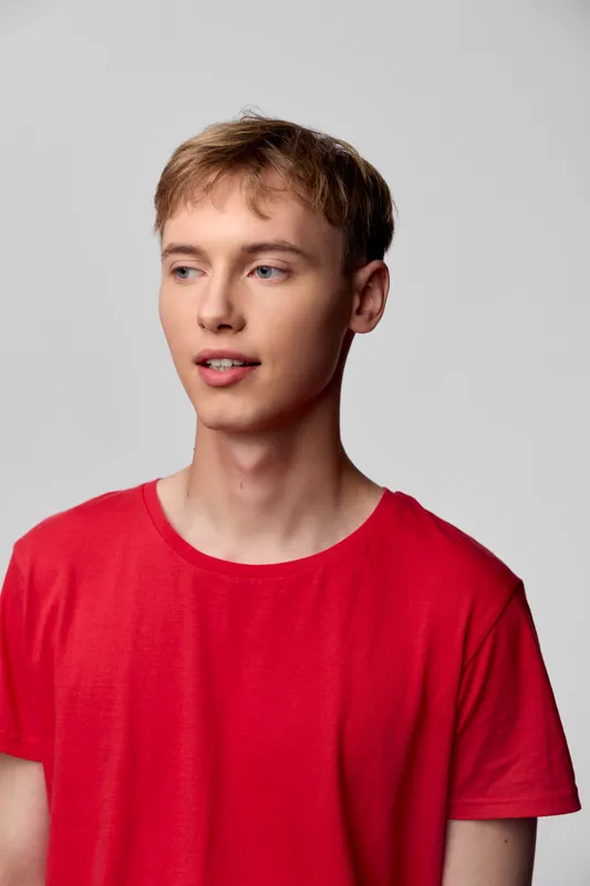 Young man in a red shirt stands in a simple studio setting with a relaxed pose and natural expression under soft studio lighting, showcasing casual style and an approachable mood