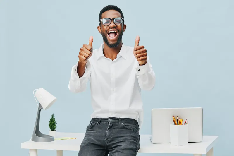 Cheerful young man wearing glasses and white shirt sitting at modern office desk smiling and giving thumbs up isolated on light blue background studio portrait. People lifestyle positivity.
