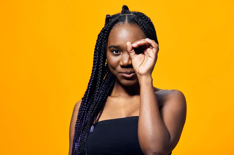 Confident young African woman playfully posing with braided hair against a bright yellow background, showcasing her self assurance and charisma with expressive gestures