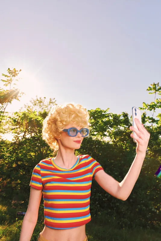 Young woman with curly blonde hair wears a colorful striped crop top and blue sunglasses as she selfies outdoors in bright sunlight, smiling with greenery in the background.