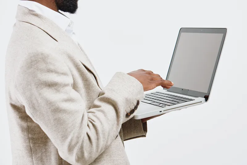 Professional woman working on laptop with blank screen, wearing beige sweater and white shirt, isolated on light grey background. Business, technology, startup, office, workplace concept.