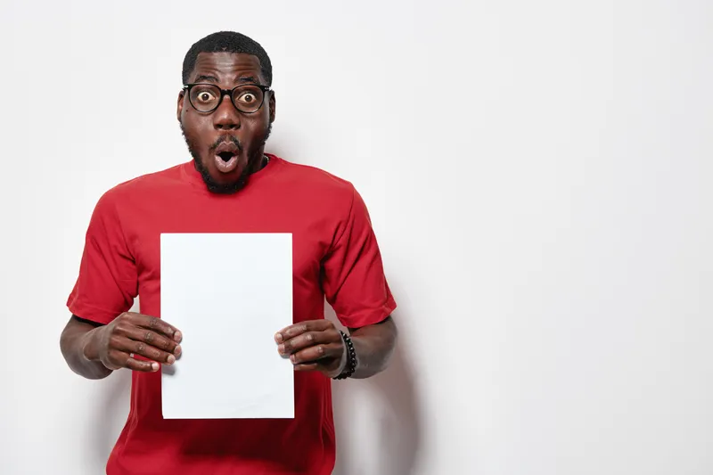 African American man with dark skin and glasses wearing red shirt holding blank white paper with surprised expression on white background for presentation or message.