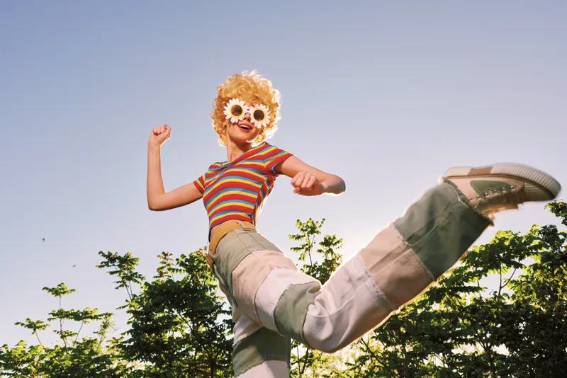 Joyful woman wearing a flower mask leaps outdoors in a striped top and wide pants, capturing a playful retro lifestyle moment against a bright sky and green trees. The scene radiates warmth and