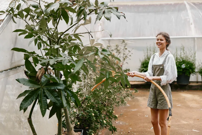 Young woman in a greenhouse watering plants, wearing casual clothing, expressing happiness amidst lush greenery and natural light