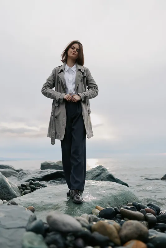 Young woman in a grey coat and dark pants stands confidently on rocky shore. Overcast sky and calm sea create a serene natural outdoor scene with casual fashion style.