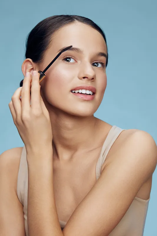 Happy young woman applying eyebrow gel, showcasing beauty and skincare routine, light background with a soft color palette, focusing on self care and makeup essentials