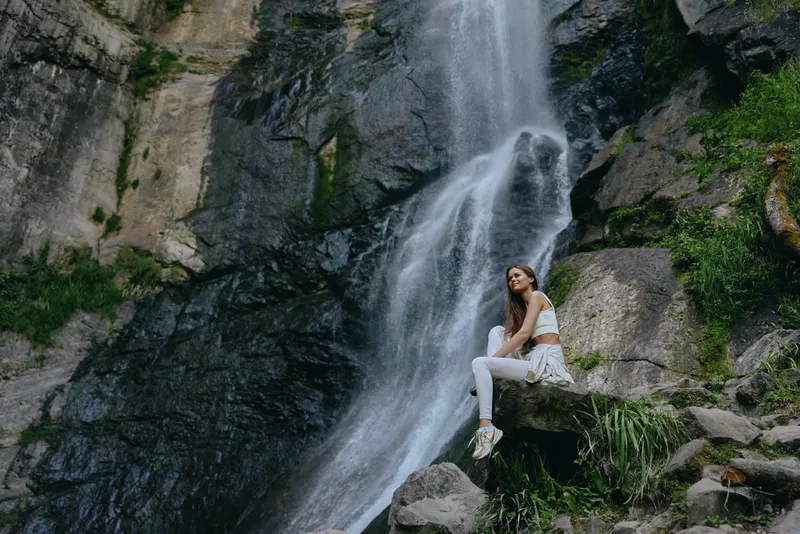 woman sitting near waterfall outdoor, surrounded by rocks and green plants, wearing white clothes and sneakers in natural environment