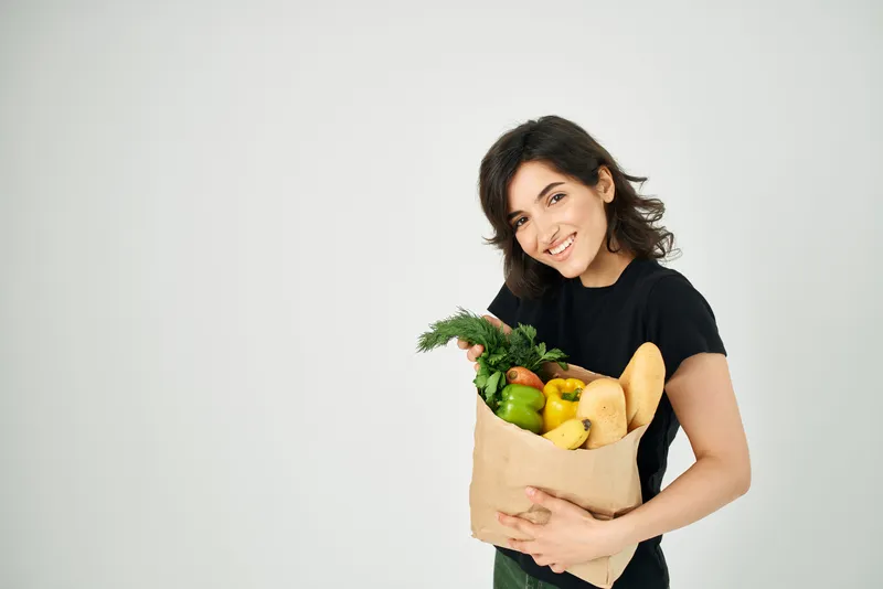 Весёлая брюнетка в чёрной футболке с пакетом продуктов из супермаркета, совершающая покупки