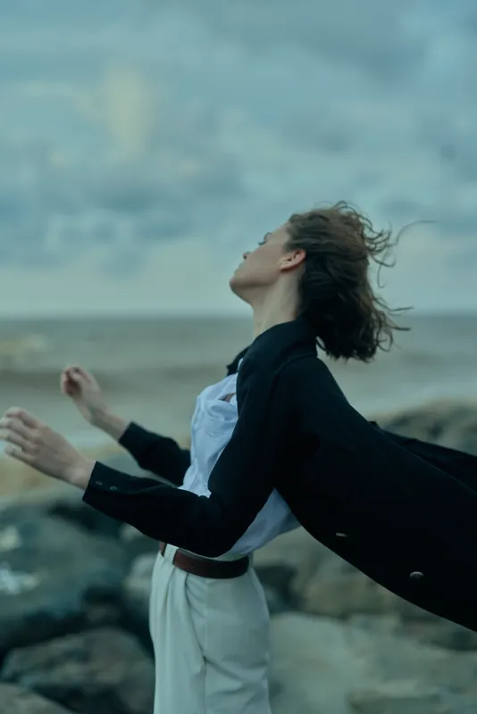 Freedom, wind, coast, flowing cape, open arms against the sea breeze, a dramatic fashion silhouette with moody light and coastal atmosphere