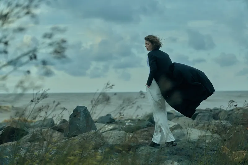 A wind swept coastal scene with a woman in a flowing coat standing on jagged rocks along the shoreline, gazing toward the ocean, a dramatic landscape of travel and freedom.