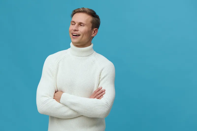 Smiling man with crossed arms wearing a cozy white sweater, standing against a vibrant blue background in a bright studio setting. Confident, happy, casual, relaxed, male, lifestyle, autumn fashion.
