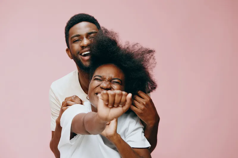Joyful couple laughing together against a pastel pink background, showcasing love, happiness, and playful emotions through their cheerful expressions