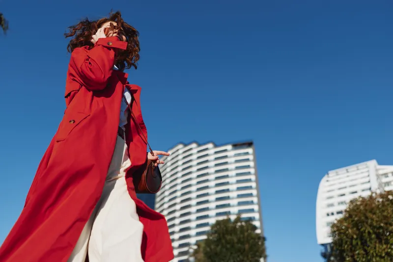 Woman in red coat walking in city with modern buildings under clear blue sky. Low angle view captures urban lifestyle and fashion concept.