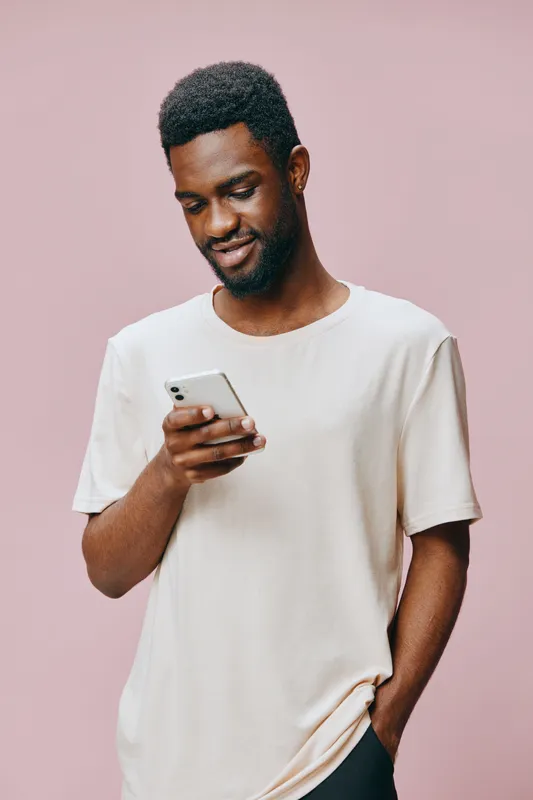 Young African man smiling while using a smartphone, wearing a casual light colored shirt against a pastel pink background, embodying modern lifestyle and happiness
