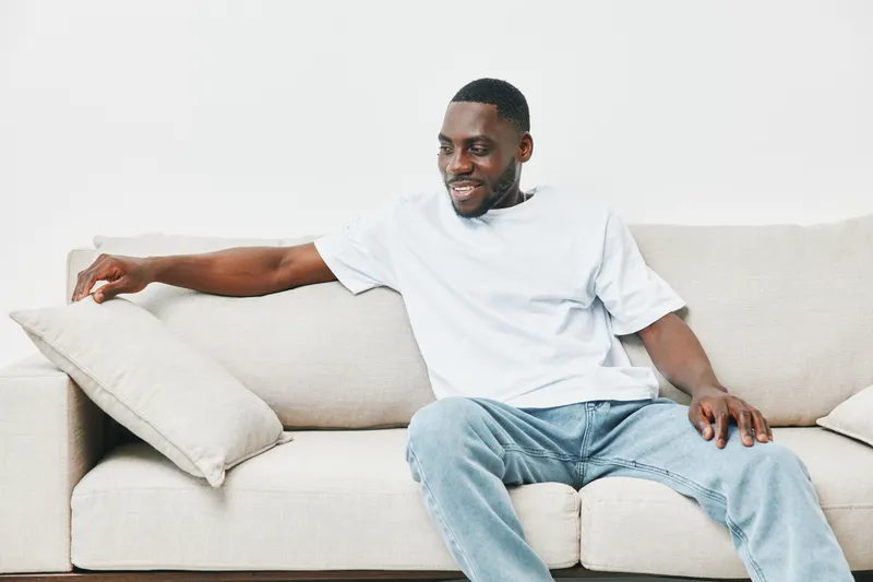 Young Black man relaxed on a sofa, wearing casual attire, smiling while adjusting a cushion in a bright living room setting