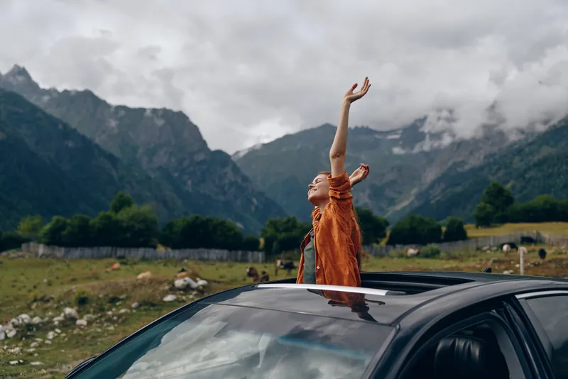 outdoor scene with a person standing through a car sunroof wearing an orange top arms raised amid mountains conveying freedom adventure and joyful travel in nature