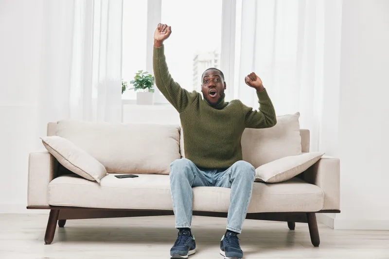 Excited young man celebrating on a couch, showcasing joy in a bright living room with soft colors and natural light, perfect for lifestyle and happiness themes