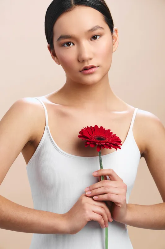 asian woman stands in a studio holding a red flower near her chest, soft lighting highlights her serene expression, creating an elegant portrait with poise, grace, and timeless appeal for lifestyle