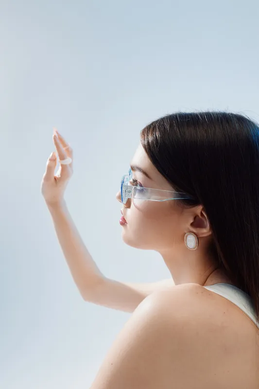 Young woman in stylish glasses and elegant earrings looks thoughtfully with raised hand. Soft lighting highlights her smooth skin and beauty. Fashion concept in minimal studio.