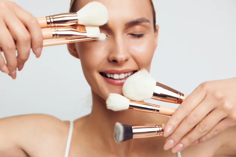 Woman applying makeup with cosmetics brush, closeup of smiling face, skincare beauty routine, hands hold brushes, soft studio light, minimal background