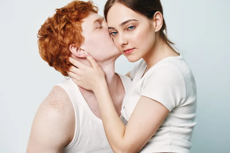 Couple in white tops with intimate pose, young man kissing woman on cheek with hands on neck and shoulder. Studio portrait with gentle emotions and close connection between people.