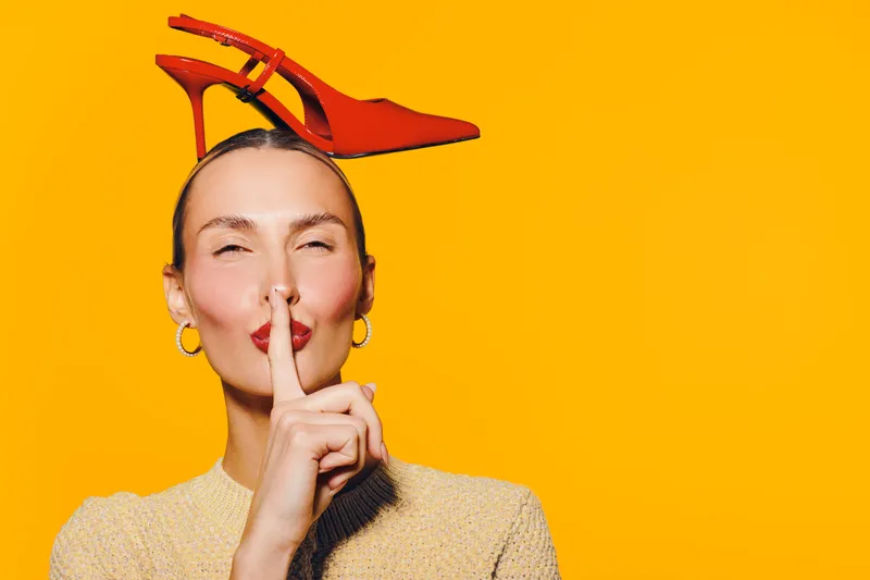 Woman with red high heel shoe on head makes shushing gesture with finger on lips against bright yellow background. Concept of silence, fashion, and playful mood.
