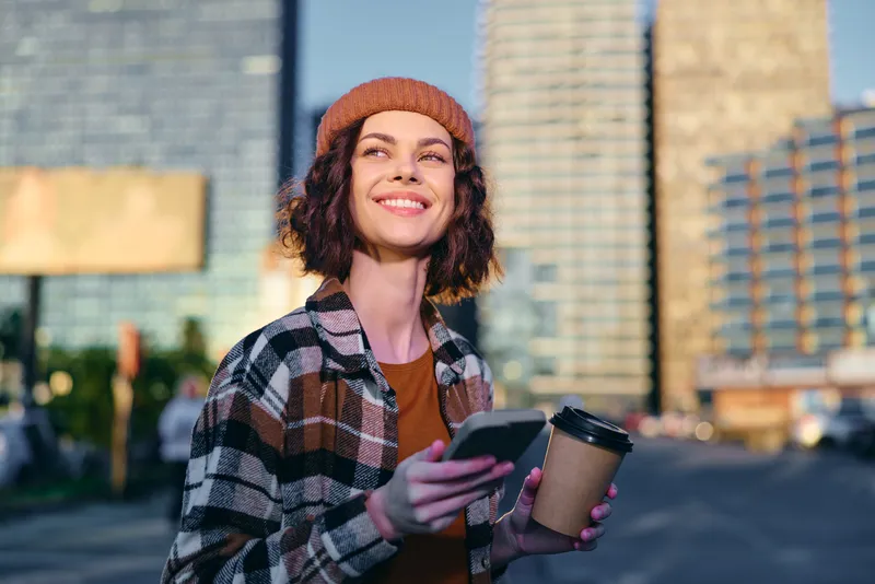 Woman holding coffee and smartphone on urban street with a smile, candid lifestyle authenticity and golden hour glow, mindful living mood and emotional storytelling in city portrait.